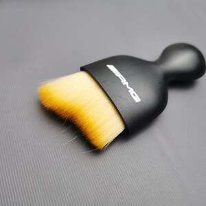 Könnte beinhalten: Eine schwarze Bürste mit rundem Griff und rechteckigem Kopf. Der Bürstenkopf hat weiche, gelbe Borsten mit orangen Spitzen. Die weißen Buchstaben "AMG" sind auf dem Bürstengriff aufgedruckt.