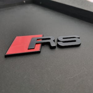 Könnte beinhalten: Schwarzes und rotes RS-Emblem. Das Emblem zeigt eine rote rechteckige Form mit den Buchstaben "RS" in Schwarz. Das Emblem ist für den Automobilbereich konzipiert und ist wahrscheinlich ein Autoabzeichen.