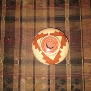 May include: A round, wooden container with a geometric design in shades of beige and orange. The container has a central, circular opening and a decorative pattern around the top edge. The item appears to be handmade.