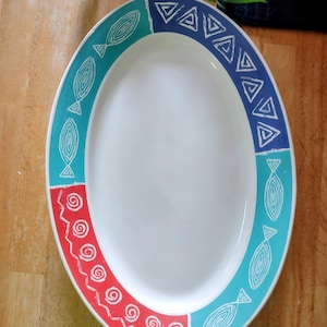 May include: Oval serving platter with a white center and a decorative border. The border features alternating sections of teal, red, and blue with white geometric and fish-like designs. The platter is on a wooden surface.