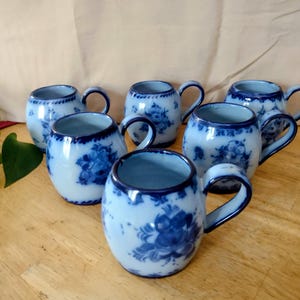 May include: Six blue and white ceramic mugs with floral designs. Each mug has a rounded shape, a dark blue rim, and a matching handle. The mugs are arranged on a light wooden surface.