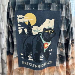 May include: Plaid button-down shirt with a large patch featuring a bear wearing sunglasses, a Colorado flag, and the text "Breckenridge CO." The shirt has a faded, distressed look with a mix of black, white, and tan colors.