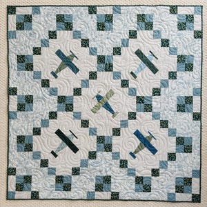 May include: A square quilt featuring a repeating pattern of blue and green squares and stylized aeroplanes. The aeroplanes are in shades of blue and green, set against a white background with a quilted swirl design.