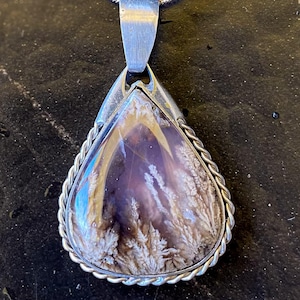 Könnte beinhalten: Eine silberne Anhängerkette mit einem tropfenförmigen Stein mit einem lila und weißen Muster. Der Stein ist von einem silbernen, seilartigen Rand umgeben und hängt an einer filigranen Silberkette. Der Anhänger ist ein einzigartiges Schmuckstück.
