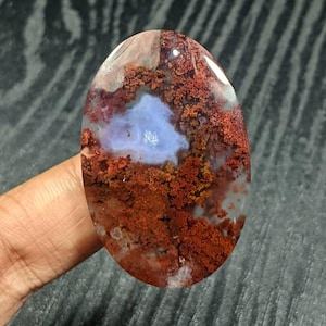 May include: An oval-shaped polished gemstone featuring a translucent light blue center surrounded by intricate, moss-like reddish-brown inclusions. The stone has a smooth, glossy surface that reflects light, showcasing a distinct pattern of organic, branching mineral formations within the clear, pale blue base.