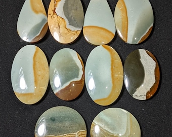 May include: A collection of polished gemstones in various shapes, including teardrop, oval, and round. The stones feature a mix of colors, including light blue, tan, brown, and gray, with a glossy finish. The gemstones are arranged on a dark surface.