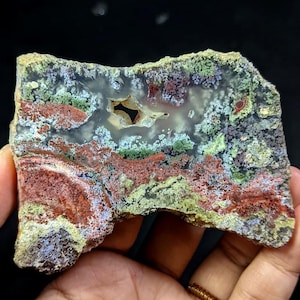 Könnte beinhalten: Eine farbenfrohe, natürliche Achatscheibe mit einer Vielzahl von Farben. Der Stein weist rote, grüne, blaue und weiße Muster mit einem zentralen Hohlraum auf. Die rauen Kanten und einzigartigen Muster machen es zu einem dekorativen Artikel.