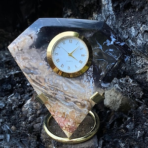 Puede incluir: Un reloj de cuarzo con bisel dorado y números romanos. El reloj está engastado en una piedra en forma de diamante con patrones negros, marrones y blancos. El reloj se apoya en un soporte dorado.