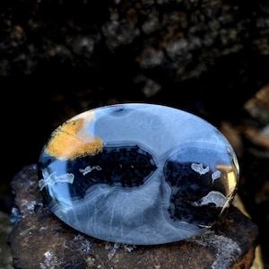 May include: Oval-shaped polished stone with a mix of colours. The stone features a gold-coloured section, a dark black area, and a light blue-grey base. The stone is resting on a rough, dark brown surface.