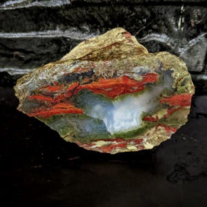 Puede incluir: Una piedra natural colorida con un patrón único. La piedra presenta capas de tonos rojos, verdes, blancos y azules. La textura rugosa y la forma irregular le dan una apariencia cruda y orgánica. Un hermoso ejemplo de arte natural.