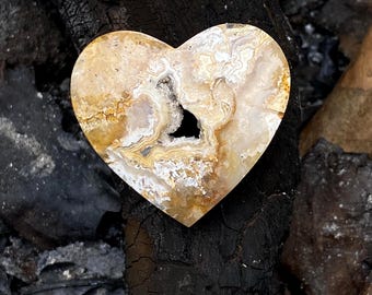 Puede incluir: Una piedra en forma de corazón con un centro de geoda. La piedra presenta una mezcla de colores beige, marrón y blanco, con un centro hueco. La piedra está colocada sobre un fondo oscuro y texturizado, creando un contraste.