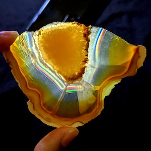 May include: A translucent, polished agate geode slice with a vibrant yellow center and a brown, textured edge. The stone displays bands of color, including orange, white, and iridescent rainbow hues, creating a striking visual effect. The geode is held by a finger.
