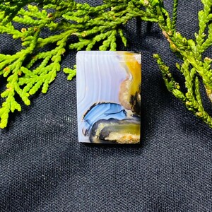 May include: A rectangular gemstone with a polished surface. The stone features a blend of colors, including white, blue, yellow, and brown, with a layered, banded pattern. The gemstone is set against a dark background with green foliage.