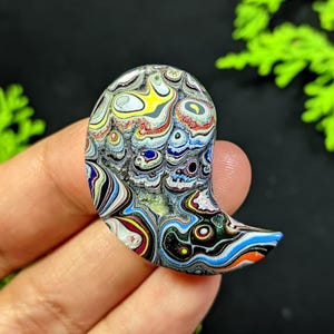 May include: A teardrop-shaped stone with a vibrant, swirling pattern of colours including yellow, blue, red, and black. The stone is held in a hand, highlighting its polished surface and intricate design. The stone's shape and pattern suggest it could be used in jewellery or as a decorative item.
