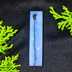 May include: A rectangular, light blue resin pendant with a dark stem and a small, detailed floral element at the top. The pendant is set against a dark background with green foliage on either side.