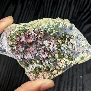 May include: A natural stone with an irregular shape and a diverse colour palette. The surface features a mix of green, white, and reddish-purple shades, with a moss-like texture. The stone is held by a hand, highlighting its unique, organic form.
