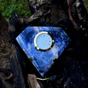 May include: A diamond-shaped clock with a blue and white stone face and a gold-colored bezel. The clock has Roman numerals and gold-colored hands. The clock is set on a dark, textured surface.