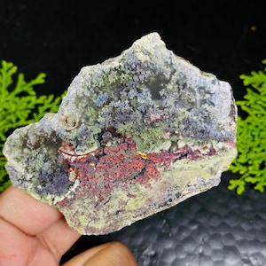 May include: A natural stone specimen with a rough, irregular shape. The surface displays a mix of colours, including red, green, blue, and grey, with a textured, moss-like appearance. The stone is held in a hand.