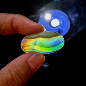 Peut inclure: Une pierre précieuse ovale et irisée tenue entre les doigts. La pierre affiche un spectre de couleurs, dont le jaune, le vert et le bleu, avec un motif rayé. Une source de lumière bleue est visible en arrière-plan, illuminant la pierre.