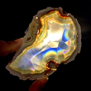 May include: A translucent, polished agate slice with a natural, irregular shape. The stone displays concentric bands of color, including yellow, orange, and blue, with a central, crystalline area. The agate is held against a black background, highlighting its vibrant hues.