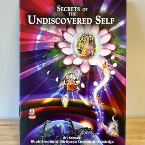 May include: Book cover with a colorful illustration of a lotus flower, a couple in the center, and a chain around a globe. The title is "Secrets of the Undiscovered Self" and the author is "Sri Srimad Bhaktivedanta Narayana Gosvami Maharaja".