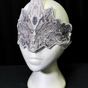 Può includere: Una corona ricamata argento e grigio con una gemma blu centrale, esposta su una testa di manichino bianca. La corona presenta intricati dettagli simili a pizzo e un design a punta. Il testo "Phantomasy" è visibile.