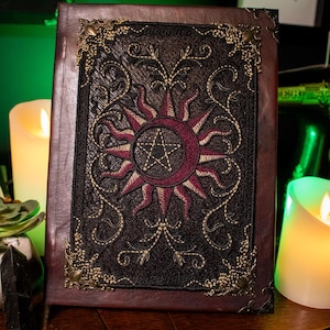 May include: A rectangular brown journal with a dark fabric center panel featuring an embroidered sun and crescent moon design with a central pentagram. The cover is accented with ornate brass-toned metal corner protectors and decorative stitching.