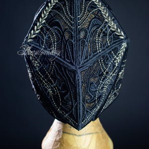 May include: A black cap with intricate embroidered detailing in a gold color. The cap is displayed on a mannequin head against a dark background. The embroidery features floral and geometric patterns.