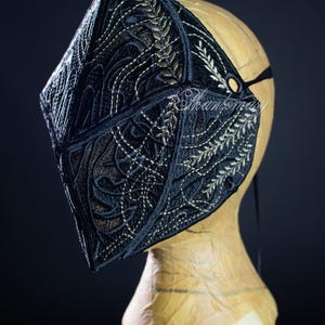 May include: Black embroidered mask with intricate gold and white floral and geometric patterns. The mask is displayed on a mannequin head, showcasing its detailed design and craftsmanship. A black strap secures the mask.