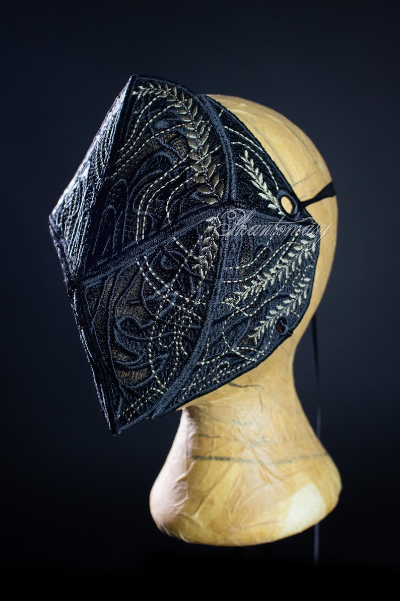 May include: Black embroidered mask with intricate gold and white floral and geometric patterns. The mask is displayed on a mannequin head, showcasing its detailed design and craftsmanship. A black strap secures the mask.
