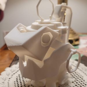 May include: A 3D-printed white sculpture of a stylized creature with a geometric design. The sculpture features a large, open mouth, a ring-shaped handle, and a series of layered cylindrical shapes. The overall aesthetic is modern and abstract.