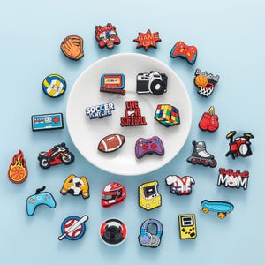 May include: A collection of colorful shoe charms featuring various designs, including sports equipment, video game controllers, cameras, and text like "Game On" and "Soccer is my life". The charms are arranged on a white plate against a light blue background.