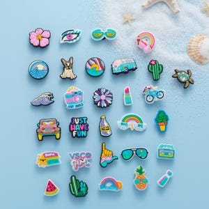 May include: A collection of colorful shoe charms with various designs, including a flower, eye, sunglasses, a dog, a wave, a camera, a lipstick, a rainbow, a cactus, a turtle, a car, a bottle, a cassette tape, a watermelon, a pineapple, and text that says "Just Have Fun", "VSCO girl", and "Summer".