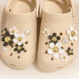 May include: A pair of beige clogs adorned with decorative flower charms in black, white, and gold. The flowers are of varying sizes and are accented with faux pearls. The clogs have multiple ventilation holes.