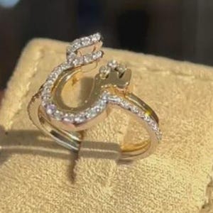 May include: A gold-toned ring with a unique design, featuring a curved shape adorned with small, sparkling gemstones. The ring is presented on a beige jewelry box, showcasing its elegant details.