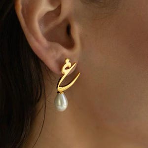 May include: Gold-toned earrings with an Arabic script design, each featuring a teardrop-shaped pearl. The earrings are worn on a person's ear, with the focus on the jewelry's elegant and minimalist aesthetic.