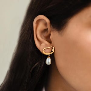 May include: A gold-colored earring with a unique design, featuring a curved top and a dangling teardrop-shaped pearl. The earring is worn on the ear, showcasing its elegant and minimalist style. The pearl is white and has a lustrous sheen.