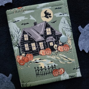 May include: A rectangular fabric wallet in sage green featuring a printed scene of a dark house, a full moon with a witch silhouette, and orange jack-o'-lanterns. The wallet is secured with a single light gray snap button closure.