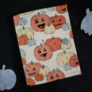 May include: A rectangular fabric wallet featuring an all-over print of orange and cream-colored jack-o'-lanterns and pumpkins on a light background. The wallet is secured with a single, solid light green snap button closure on the front flap.