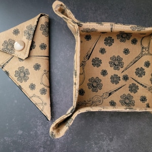 May include: A tan fabric tray with four snap-button corners and a matching triangular pouch with a single snap closure. Both items feature a repeating black print of small flowers and vintage-style sewing scissors on a light brown background.