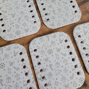 May include: Five light gray, rectangular pieces with rounded corners, each featuring a repeating pattern of Christmas-themed line art. The designs include Santa hats, snowflakes, and candy canes. Each piece has multiple holes.