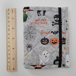 May include: A gray quilted Halloween-themed notebook cover with a black elastic closure. The cover features a spiderweb, pumpkins, ghosts, and the text "DRINK IF YOU DARE" and "Trick". The notebook measures approximately 5 inches tall.