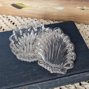 May include: Two clear, glass seashell-shaped dishes. The dishes are resting on a dark blue book. The dishes have a detailed, textured design. The background includes a painting of white flowers in a dark vase.