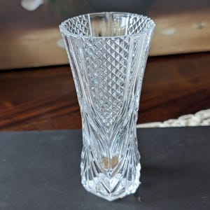 May include: A clear, faceted glass vase with a diamond pattern. The vase has a flared top and a geometric base. The glass is transparent, allowing light to pass through. The vase is likely for displaying flowers or as a decorative item.
