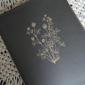 May include: A rectangular black binder featuring a gold-colored line art illustration of a clover plant with multiple stems, leaves, and flower heads centered on the front cover. The binder has a smooth, matte finish and a clean, structured design.