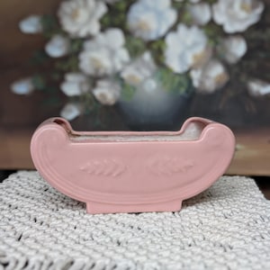 May include: A pink ceramic planter with a curved design and embossed leaf details. The planter sits on a white macrame surface, with a blurred painting of white flowers in the background. The planter is a soft, pastel pink.