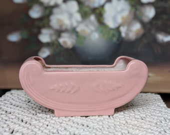 May include: A pink ceramic planter with a curved design and embossed leaf details. The planter sits on a white macrame surface, with a blurred painting of white flowers in the background. The planter is a soft, pastel pink.