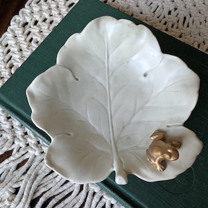 May include: A white ceramic trinket dish shaped like a large leaf with a small, metallic gold-colored frog figurine attached to the lower right side. The dish features embossed vein details across its surface and has a smooth, matte finish.