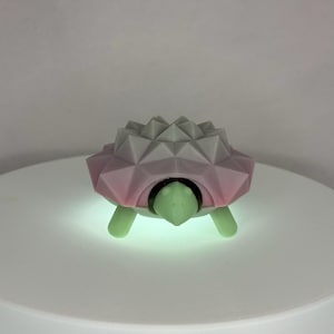 May include: A geometric turtle figurine with a pink and gray shell, green head and legs. The shell has a faceted design. The turtle is on a white surface.