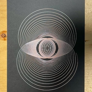 May include: A black print featuring a geometric design of concentric circles and an eye-like shape in the center. The lines are a light pink color, creating a contrast against the dark background. The artwork has a minimalist aesthetic.